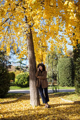 Frau, die im Herbst unter einem Baum steht und mit ihrem Handy telefoniert - MRRF00734