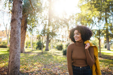 Smiling friend holding jacket on shoulder while standing at park - MRRF00739