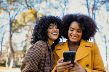 Freunde lächelnd beim Benutzen eines Smartphones im Park stehend - MRRF00743