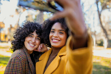 Smiling woman taking selfie with friend through smart phone while standing at park - MRRF00744