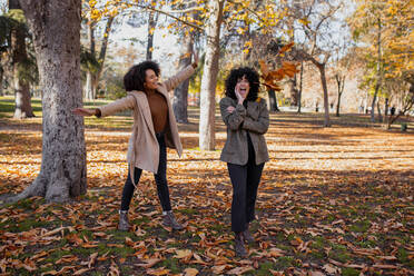 Cheerful friend throwing leaf on woman standing with hand on chin at park - MRRF00757