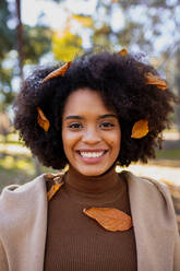 Curly hair woman with dry leaf on hair smiling while standing at park - MRRF00758