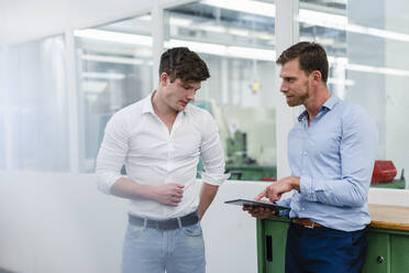 Männlicher Ingenieur schaut auf ein digitales Tablet, während er sich mit einem Geschäftsmann in einer Fabrik unterhält - DIGF13791