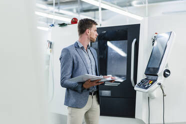 Male entrepreneur holding file document while looking at machinery equipment in industry - DIGF13809