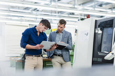 Male coworkers looking at file document while working by machinery in factory - DIGF13810