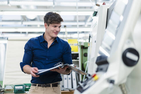 Lächelnder männlicher Ingenieur, der ein Dokument in der Hand hält, während er eine Fertigungsanlage in einer Fabrik untersucht - DIGF13819