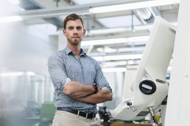 Selbstbewusster männlicher Ingenieur mit verschränkten Armen, der neben einer Produktionsanlage in einer Fabrik steht - DIGF13828