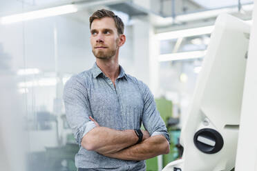 Male entrepreneur with arms crossed looking away while standing by machine in factory - DIGF13830