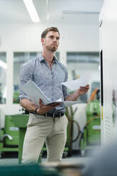 Businessman with file document day dreaming while standing by machine in factory - DIGF13845