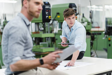 Männlicher Ingenieur, der ein digitales Tablet hält, während ein Unternehmer bei einer Besprechung in einer Fabrik wegschaut - DIGF13849