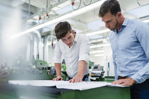 Männlicher Unternehmer mit einem Kollegen, der eine Blaupause auf dem Schreibtisch in einer Fabrik betrachtet - DIGF13877