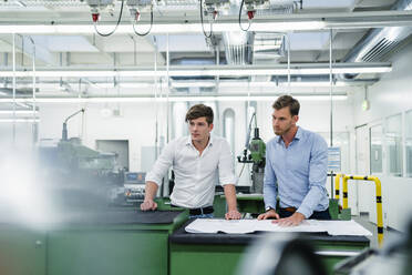 Male engineers with floor plan looking at manufacturing equipment while having meeting in factory - DIGF13879