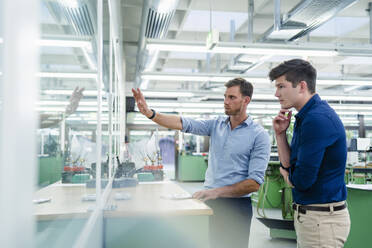 Männliche Kollegen schauen durch ein Glas, während sie in einer Fabrik diskutieren - DIGF13886