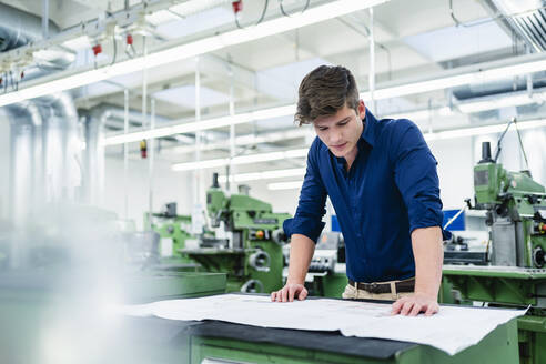 Männlicher Unternehmer, der einen Grundriss betrachtet, während er am Schreibtisch in einer Fabrik arbeitet - DIGF13890