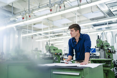 Männlicher Ingenieur mit Grundriss und Maschinenteil, der in einer Fabrik wegschaut - DIGF13892