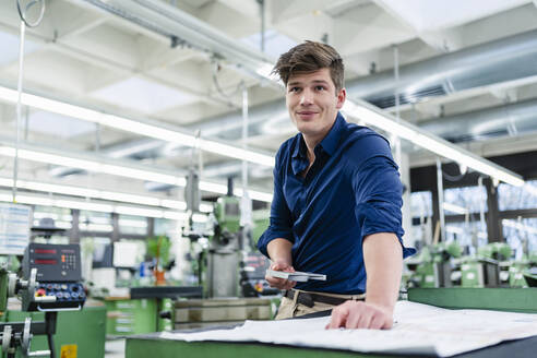 Männlicher Fachmann mit Maschinenteil, der wegschaut, während er an einem Grundriss in einer Fabrik arbeitet - DIGF13895