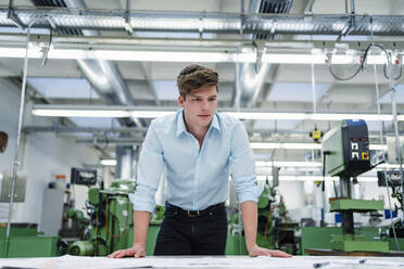 Young male entrepreneur examining blueprint at desk in factory - DIGF13911