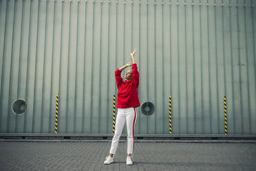 Carefree woman with hand raised standing on footpath against wall - GUSF04832