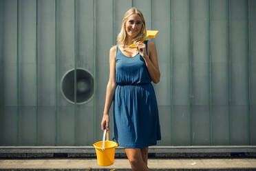Smiling woman with sand bucket and shovel standing on footpath against wall - GUSF04866
