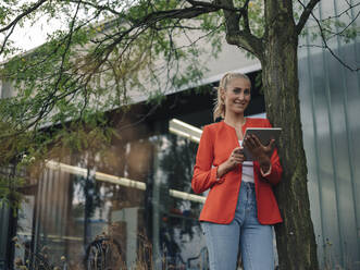 Lächelnde Geschäftsfrau mit digitalem Tablet, die vor einem Bürogebäude an einem Baum steht - GUSF04913