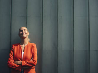 Smiling businesswoman with arms crossed against gray wall on sunny day - GUSF04920