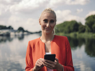 Selbstbewusste Geschäftsfrau mit Smartphone am See an einem sonnigen Tag - GUSF04928