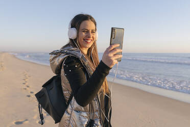 Lächelnde Frau, die ein Selfie mit ihrem Smartphone macht, während sie am Strand gegen den klaren Himmel steht - JRVF00081