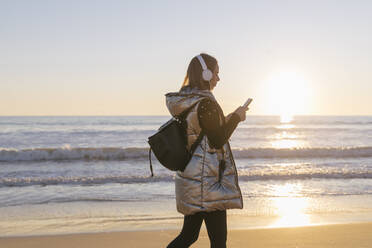 Frau, die Musik hört und ihr Smartphone benutzt, während sie am Strand bei Sonnenuntergang spazieren geht - JRVF00083