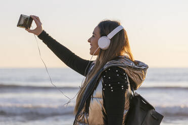 Frau nimmt Selfie beim Hören von Musik gegen das Meer bei Sonnenuntergang - JRVF00085