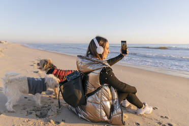 Mid erwachsene Frau, die selfie beim Sitzen mit Hunden am Strand während des Sonnenuntergangs - JRVF00087