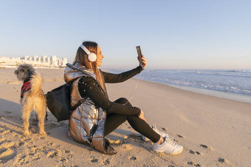 Frau, die ein Selfie macht, während sie mit ihrem Hund am Strand gegen den klaren Himmel bei Sonnenuntergang sitzt - JRVF00088