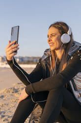 Lächelnde schöne Frau nimmt Selfie mit Handy am Strand gegen klaren Himmel - JRVF00089