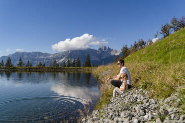 Vater sitzt mit kleiner Tochter am Ufer eines Alpensees - DIGF13946