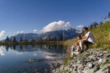 Family with little daughter sitting together on shore of alpine lake - DIGF13947