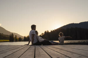 Family with little daughter sitting together at end of lakeshore jetty at sunset - DIGF13951