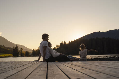 Familie mit kleiner Tochter sitzt bei Sonnenuntergang am Ende eines Stegs am Seeufer zusammen - DIGF13951