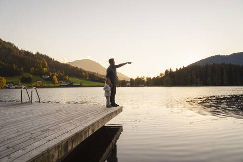 Vater und kleine Tochter stehen bei Sonnenuntergang gemeinsam am Ufer des Seestegs - DIGF13952