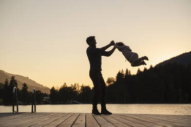Silhouetten von Vater und kleiner Tochter, die in der Abenddämmerung am Ende eines Stegs am Seeufer spielen - DIGF13955