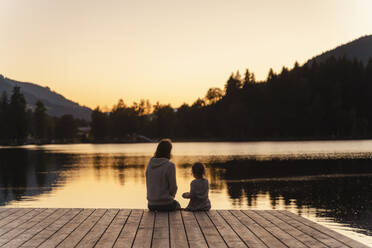 Mutter und kleine Tochter sitzen in der Abenddämmerung zusammen am Ende eines Stegs am Seeufer - DIGF13962
