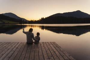 Mother and little daughter sitting together at end of lakeshore jetty at dusk - DIGF13964