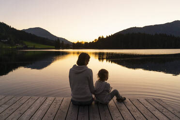 Mother and little daughter sitting together at end of lakeshore jetty at dusk - DIGF13965