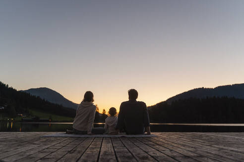 Family with little daughter sitting together at end of lakeshore jetty at dusk - DIGF13966