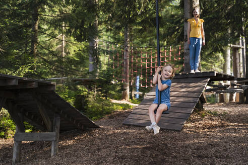 Kleines Mädchen reitet auf einer Seilrutsche im Wald, die Mutter steht im Hintergrund - DIGF13985