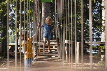 Mother assisting little daughter walking across small suspension bridge on forest obstacle course - DIGF13987