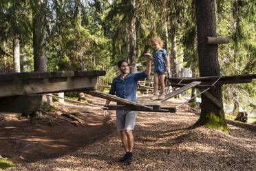 Vater hilft seiner kleinen Tochter beim Gehen über eine kleine Hängebrücke auf einem Hindernisparcours im Wald - DIGF13988