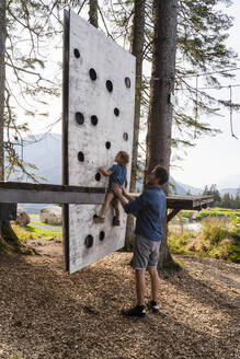 Father assisting little daughter trying to climb on top of small forest climbing wall - DIGF13989