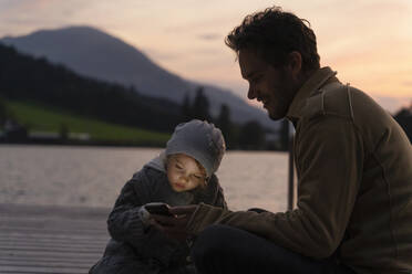 Vater und kleine Tochter spielen in der Abenddämmerung mit ihrem Smartphone auf einem Steg am Seeufer - DIGF13993