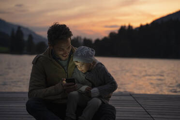 Father and little daughter playing with smart phone on lakeshore jetty at dusk - DIGF13994