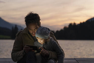 Vater und kleine Tochter spielen in der Abenddämmerung mit ihrem Smartphone auf einem Steg am Seeufer - DIGF13995
