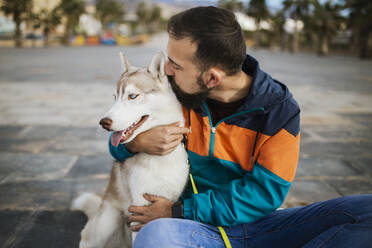 Bearded man kissing Siberian Husky while sitting on street - MIMFF00381